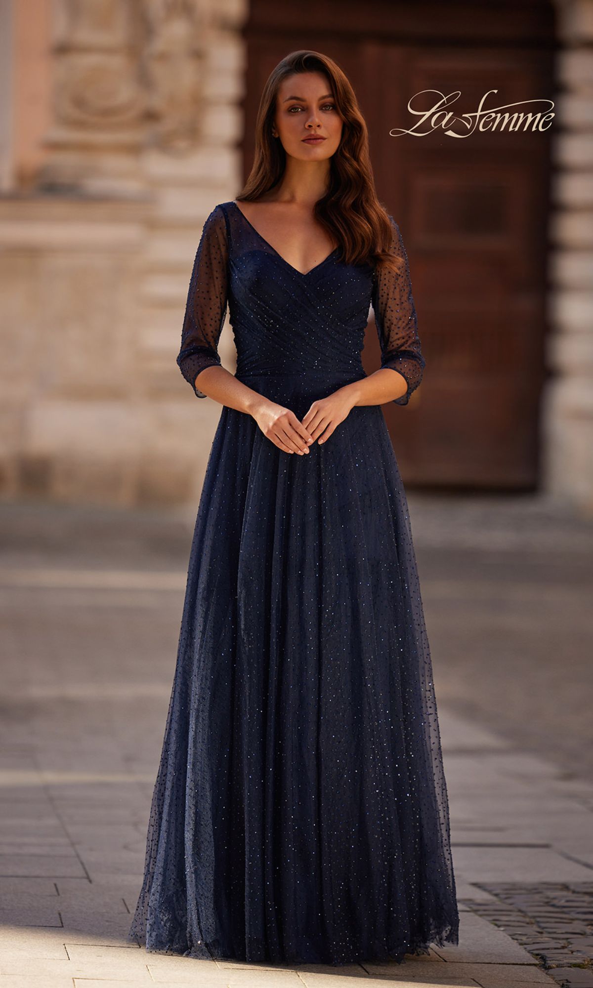 A woman stands outdoors by a stone building in the La Femme 32460 Sparkly Sleeved Tulle Formal Dress—navy blue, V-neck, with sheer three-quarter sleeves and subtle glitter.